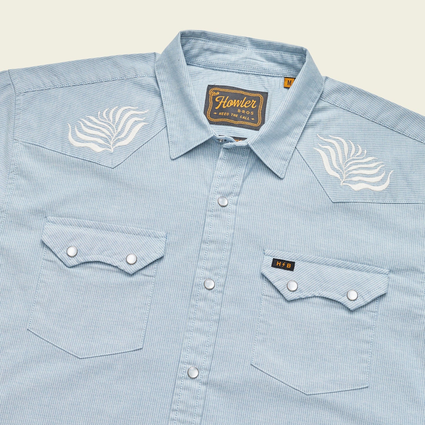 A light blue, striped Crosscut Deluxe Shortsleeve pearl snap shirt with pointed pockets and white motif embroidery on the shoulders. The shirt features a Howler brand tag on the inside collar.