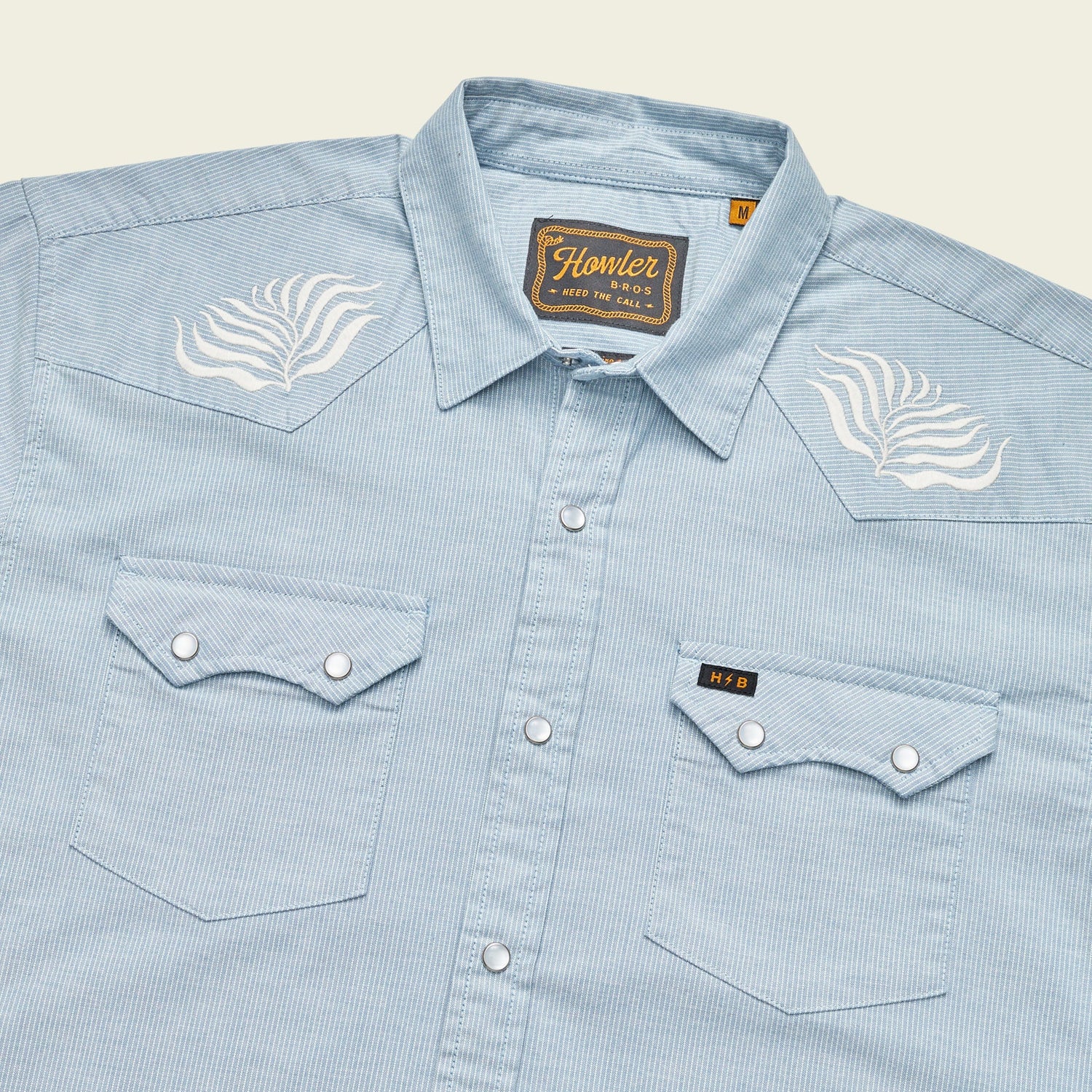 A light blue, striped Crosscut Deluxe Shortsleeve pearl snap shirt with pointed pockets and white motif embroidery on the shoulders. The shirt features a Howler brand tag on the inside collar.
