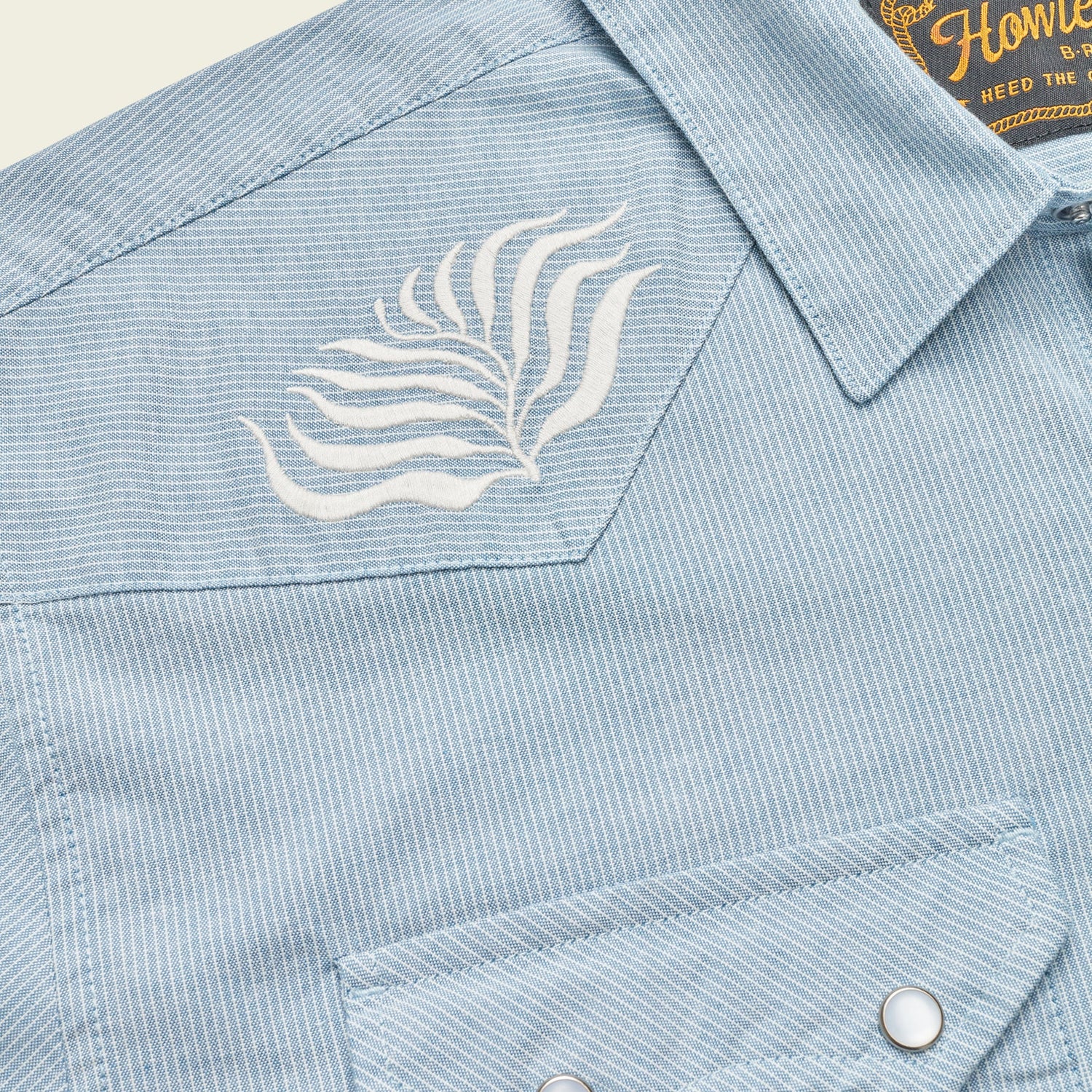 Close-up of a light blue, striped Crosscut Deluxe Shortsleeve pearl snap shirt, featuring white motif embroidery in leaf patterns on the chest.