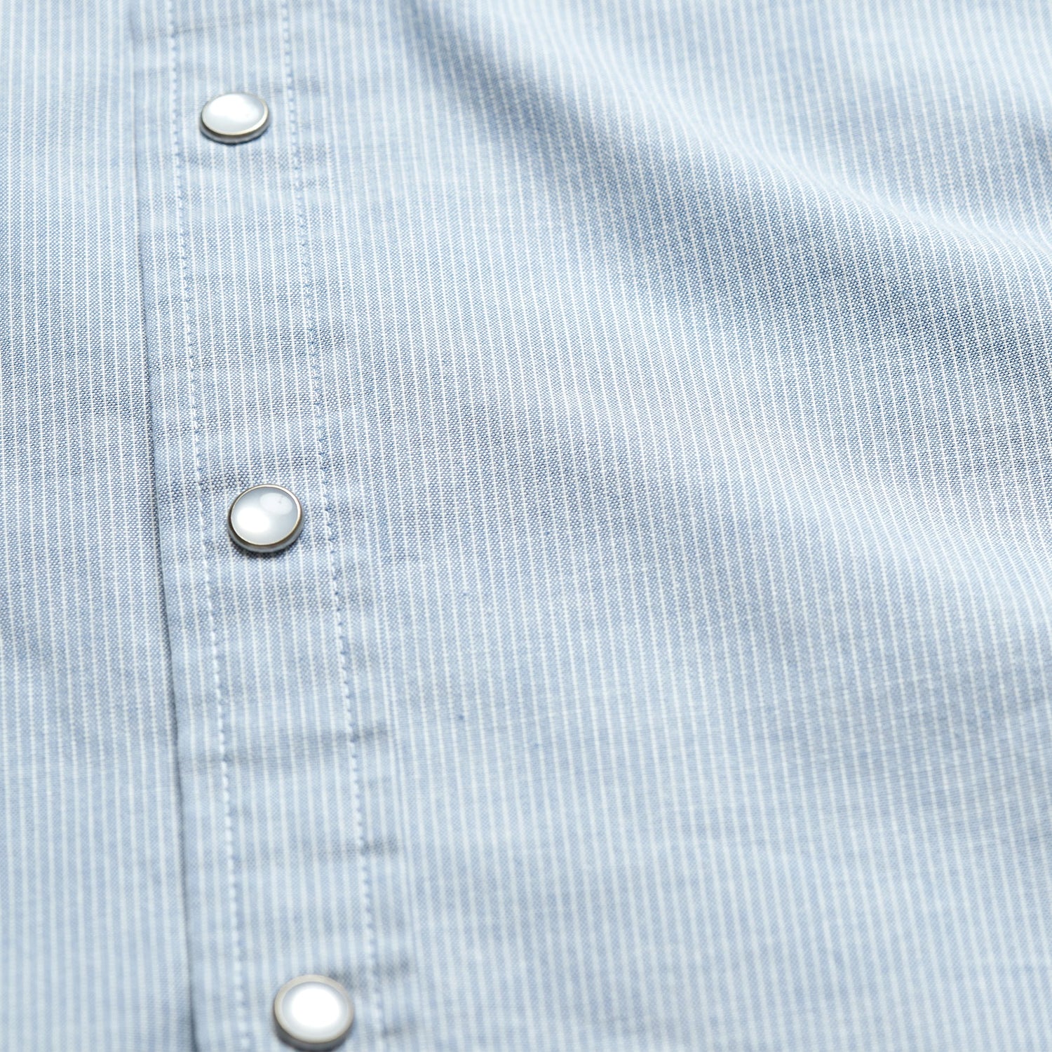 Close-up of a light blue pearl snap shirt with thin white vertical stripes and silver snap buttons along the front placket. The fabric texture, stitching details, and subtle motif embroidery are clearly visible.