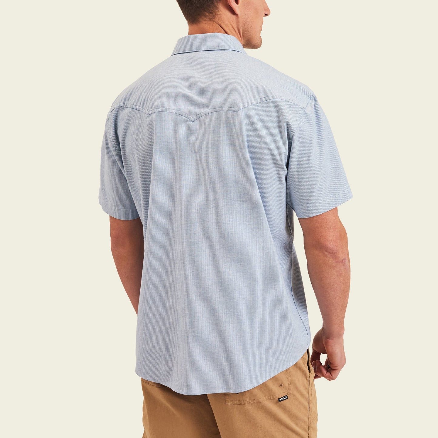 A man wearing a light blue Crosscut Deluxe Shortsleeve pearl snap shirt and tan shorts stands facing away, showing the back of the shirt with motif embroidery against a plain light background.