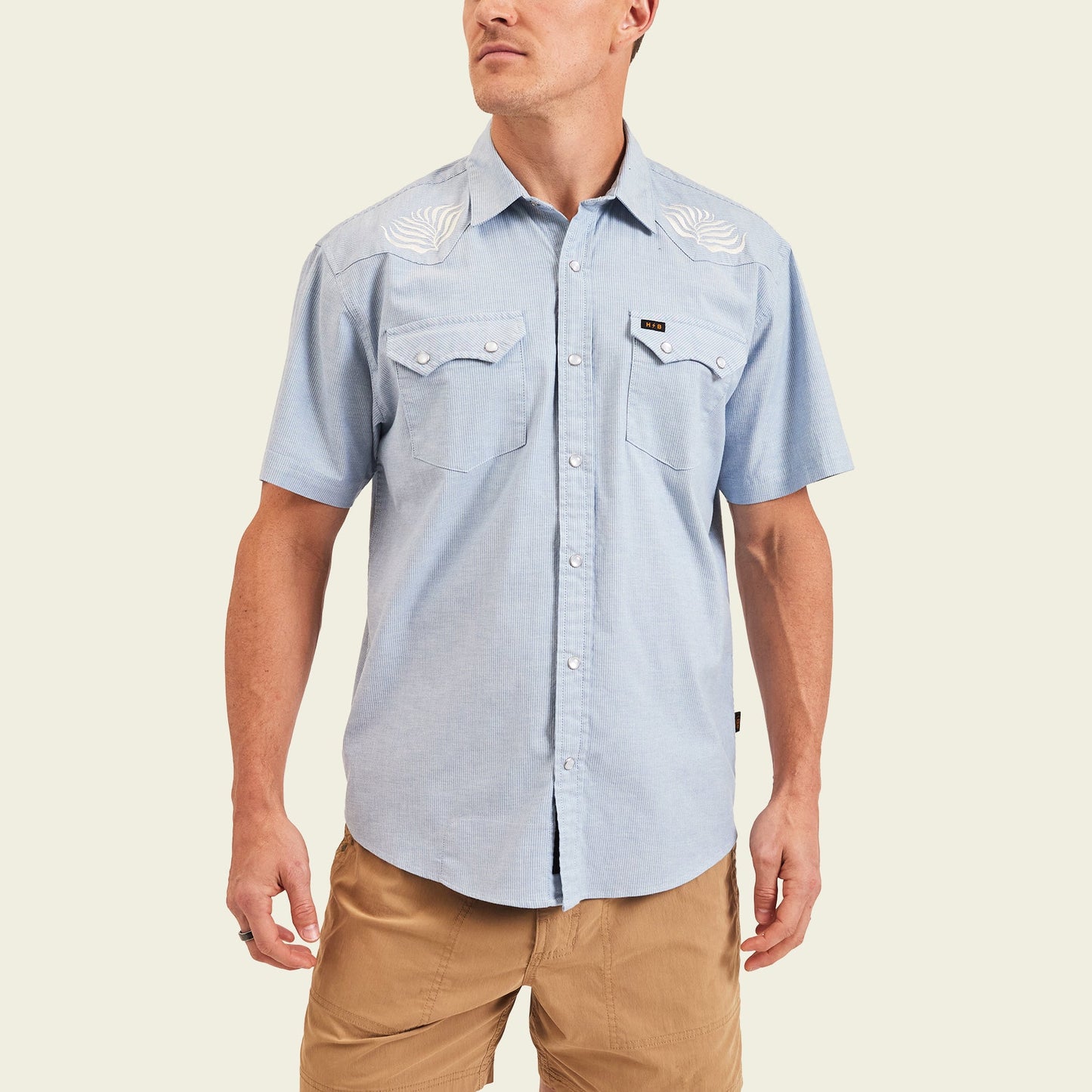 A man wears a light blue Crosscut Deluxe Shortsleeve pearl snap shirt with white motif embroidery on the shoulders and tan shorts, standing against a plain light background.
