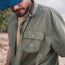 A man wearing a blue cap and the Forager Utility Shirt—a tech shirt with a mesh pocket and Howler Brothers logo—stands outdoors, looking down, ready to explore hiking trails.