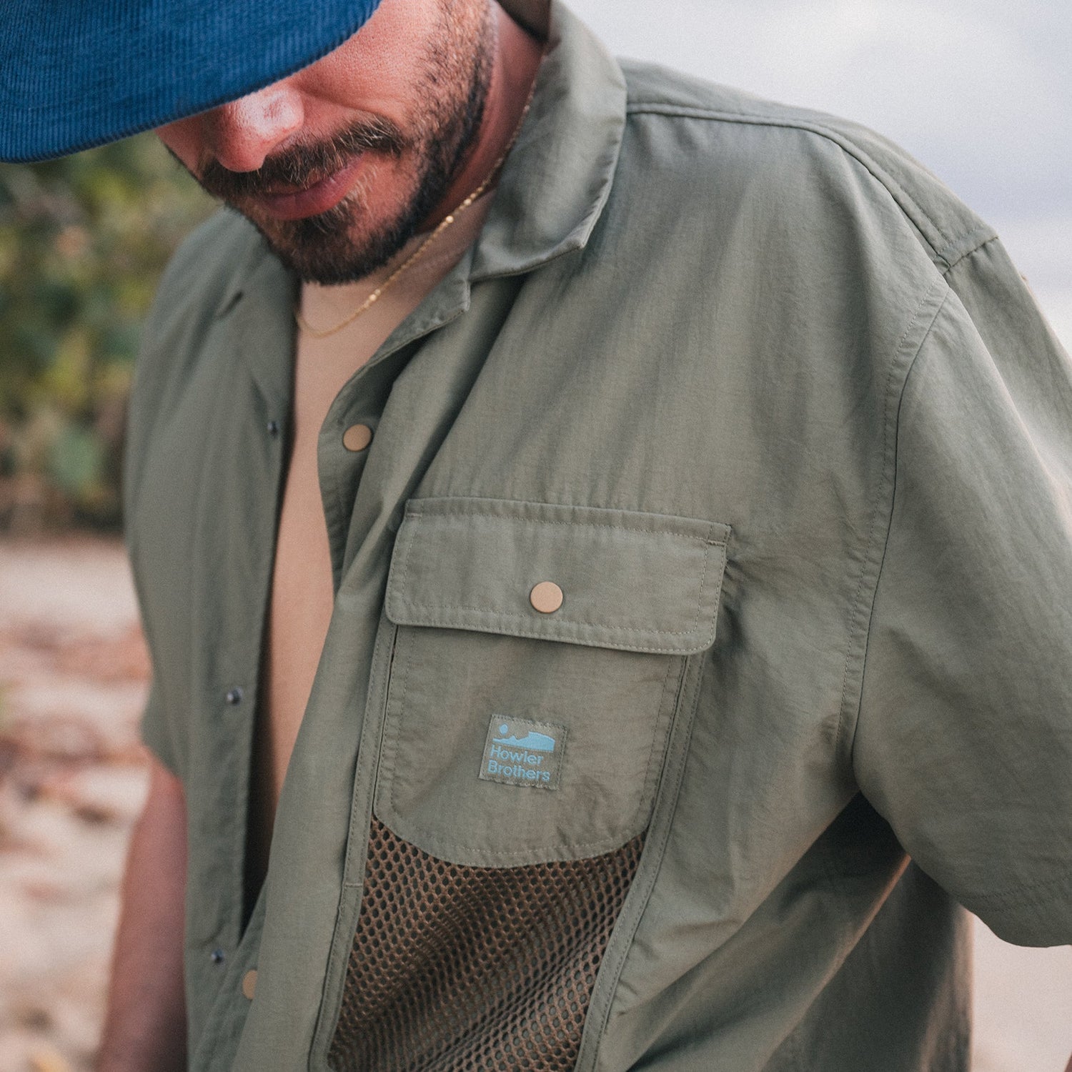 A man wearing a blue cap and the Forager Utility Shirt—a tech shirt with a mesh pocket and Howler Brothers logo—stands outdoors, looking down, ready to explore hiking trails.