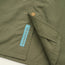 Close-up of an olive green fabric with a brass snap button and a blue Howler Brothers tag, likely part of the Forager Utility Shirt—perfect for hiking trails, featuring visible seams and a pocket for added utility.