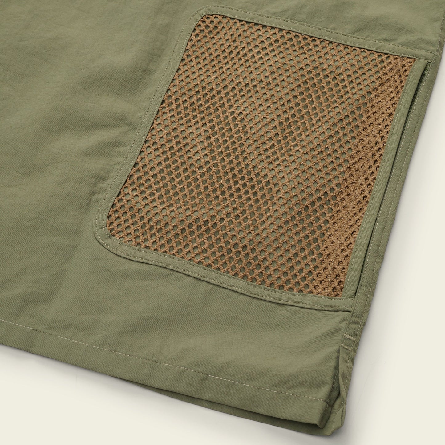 Close-up of an olive green Forager Utility Shirt fabric with a brown mesh pocket sewn on top, showing the texture and stitching details—perfect for comfort and storage on hiking trails. The edge of the fabric is visible in the bottom right corner.