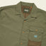 Olive green Forager Utility Shirt with short sleeves, collar, snap buttons, mesh lining, and a Howler Brothers logo patch. This tech shirt offers casual, utilitarian style—perfect for hiking trails or laid-back days out.