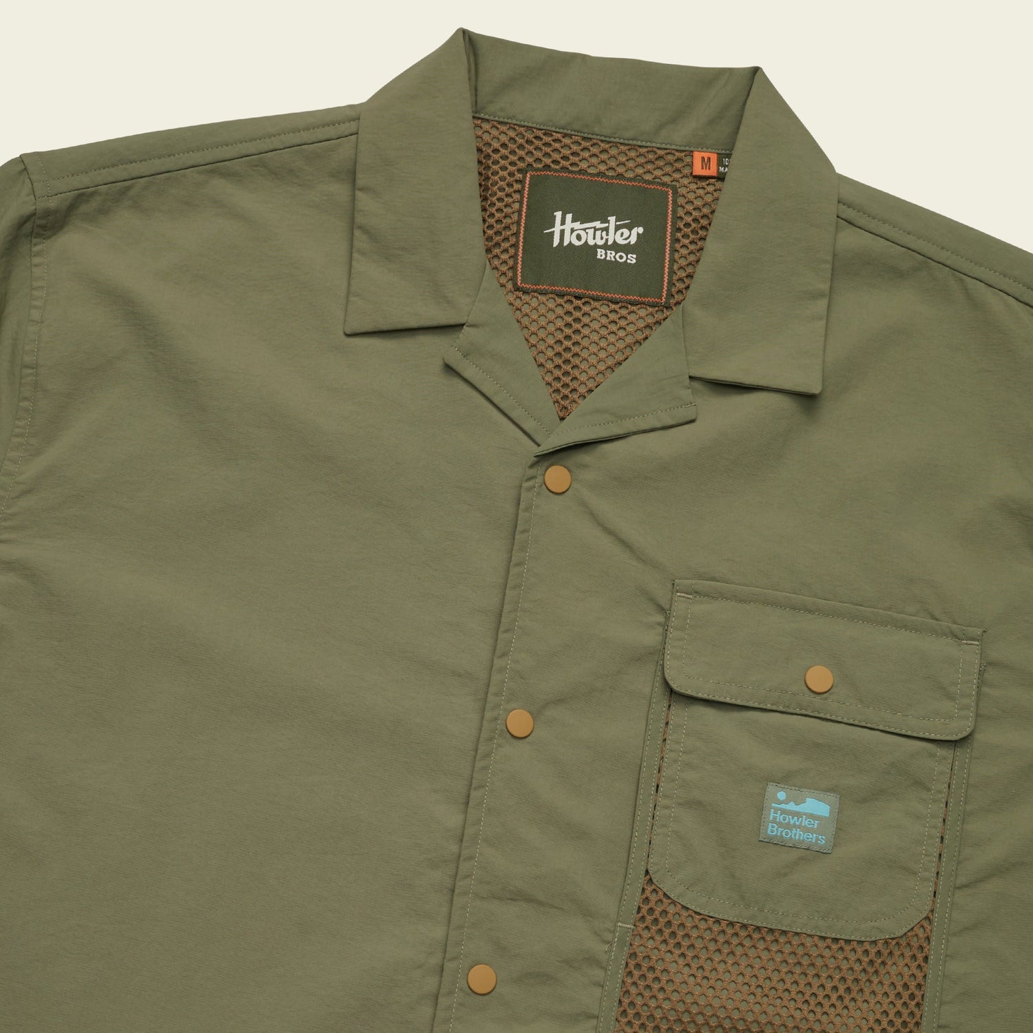 Olive green Forager Utility Shirt with short sleeves, collar, snap buttons, mesh lining, and a Howler Brothers logo patch. This tech shirt offers casual, utilitarian style—perfect for hiking trails or laid-back days out.