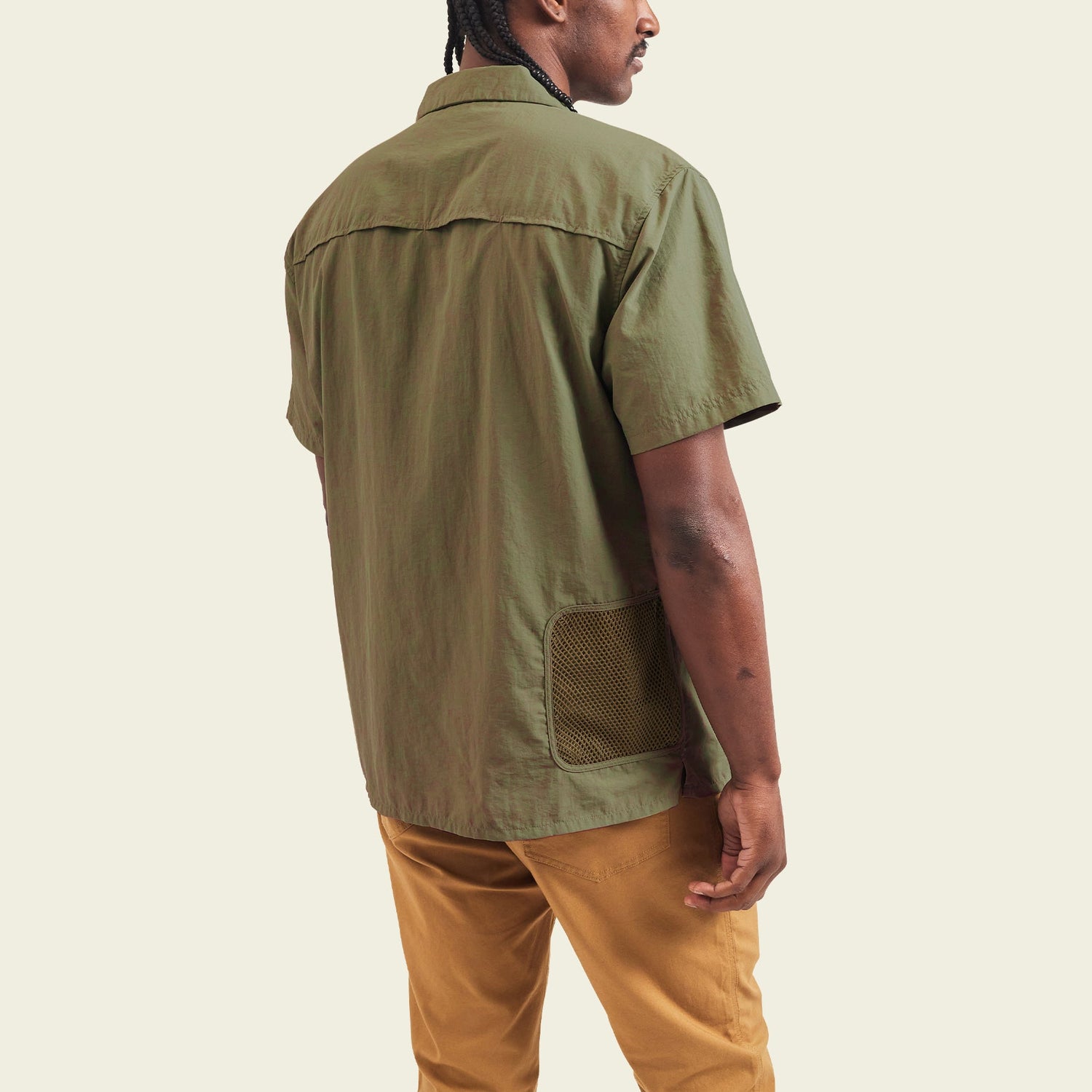 A man stands facing away, wearing the olive-green Forager Utility Shirt with mesh pocket detail—an ideal tech shirt for hiking trails—paired with tan pants, against a plain light background.