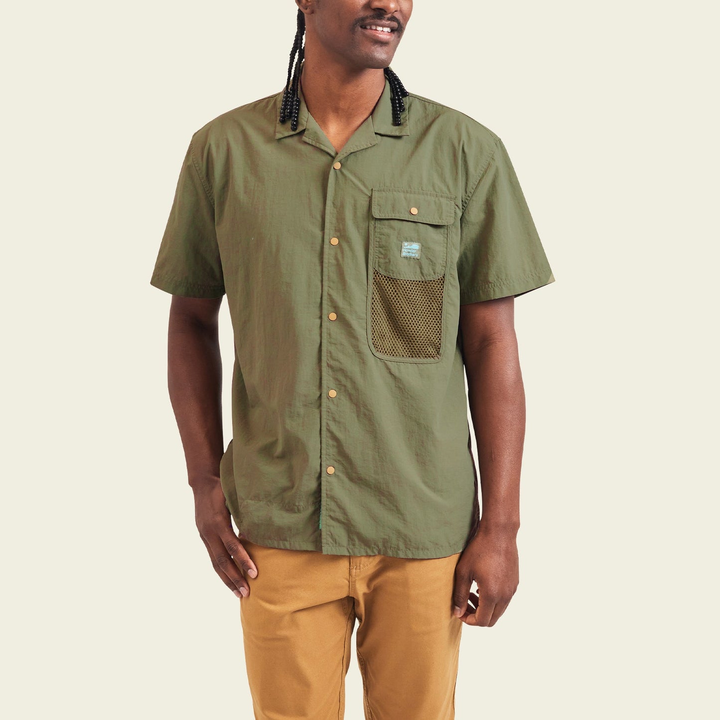 A person wearing a Forager Utility Shirt—an olive green, short-sleeve button-up with a mesh pocket—paired with tan pants, stands against a plain light background, ready for hiking trails.