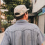 A man in a light blue Guayabera with unique embroidery and a patterned cap walks down a sidewalk, facing away from the camera. Trees, buildings, and a closed storefront appear in the background.