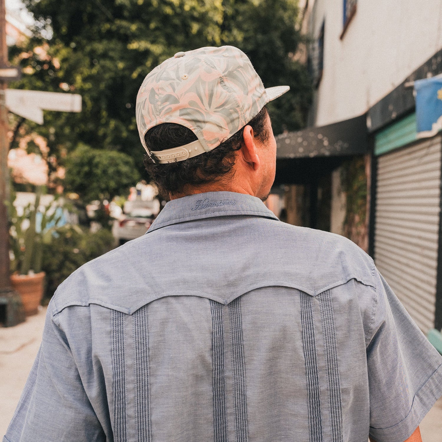 A man in a light blue Guayabera with unique embroidery and a patterned cap walks down a sidewalk, facing away from the camera. Trees, buildings, and a closed storefront appear in the background.