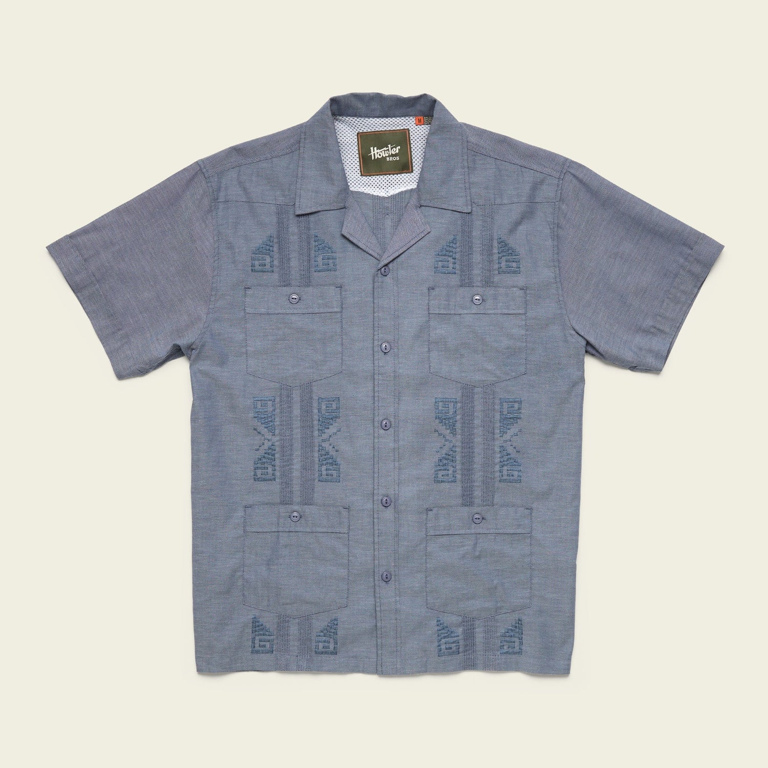 A short-sleeve, button-up blue Guayabera shirt with a subtle geometric pattern, two chest pockets, and unique embroidery, crafted from technical cotton-poly stretch fabric, displayed flat on a neutral background.