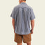 A man stands facing away from the camera, wearing a short-sleeve blue patterned Guayabera shirt with unique embroidery and brown shorts, set against a plain light background.