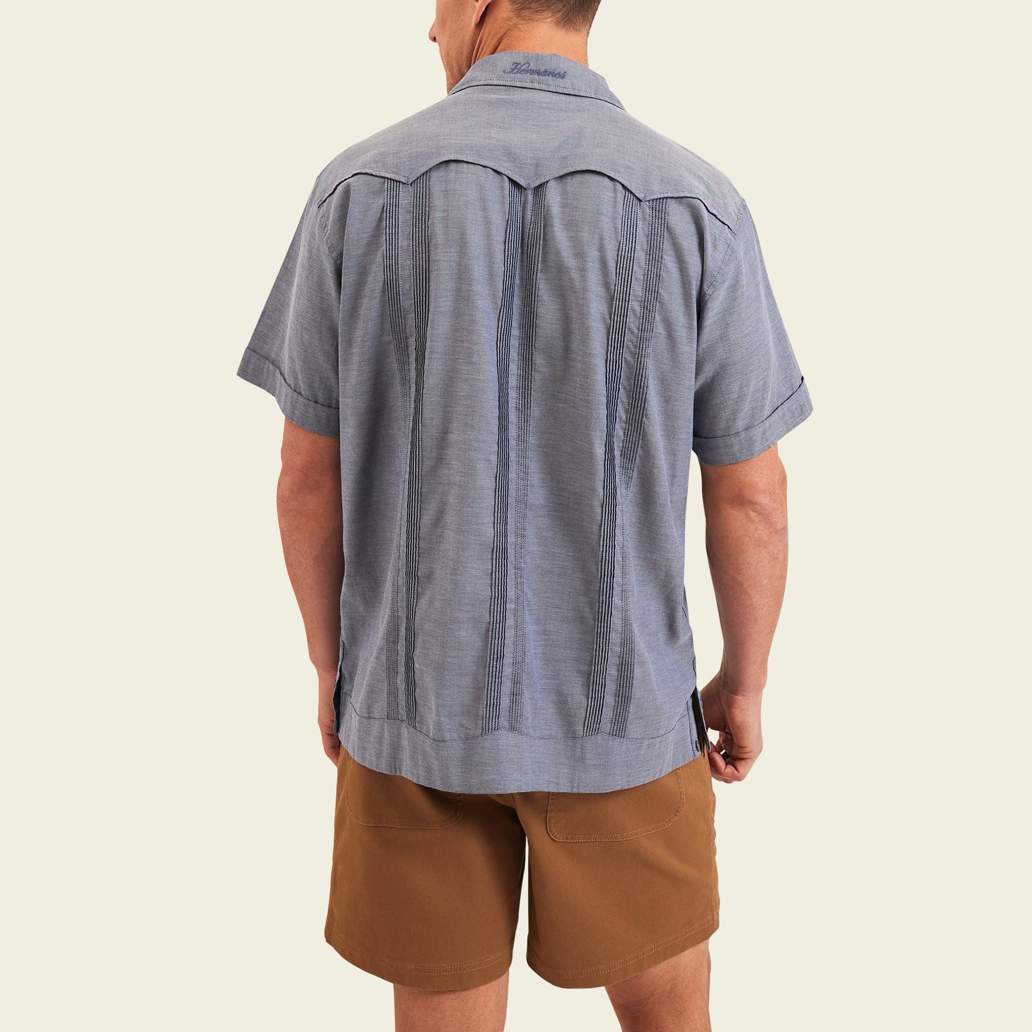 A man stands facing away from the camera, wearing a short-sleeve blue patterned Guayabera shirt with unique embroidery and brown shorts, set against a plain light background.