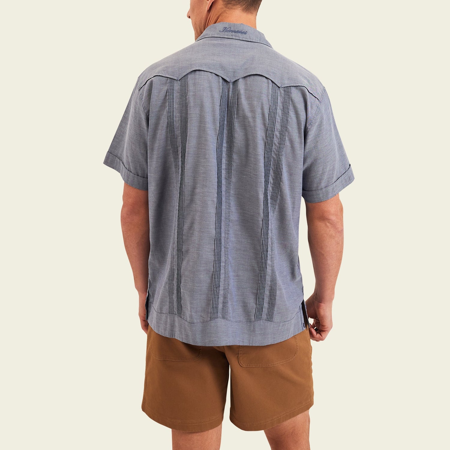 A man stands facing away from the camera, wearing a short-sleeve blue patterned Guayabera shirt with unique embroidery and brown shorts, set against a plain light background.