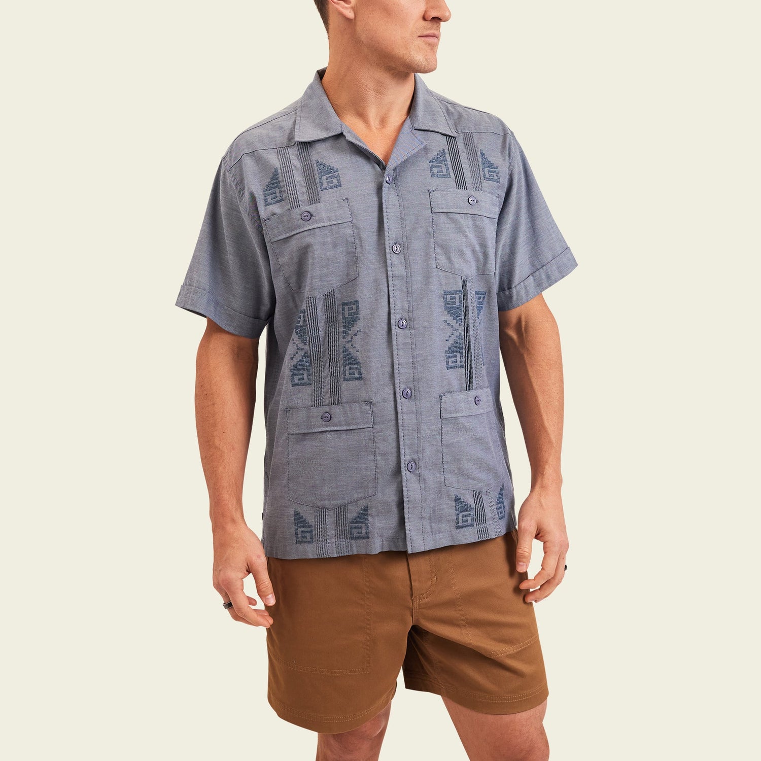 A person wearing a short-sleeve, button-up blue patterned Guayabera with unique embroidery and brown shorts stands against a plain, light background, looking off to the side.