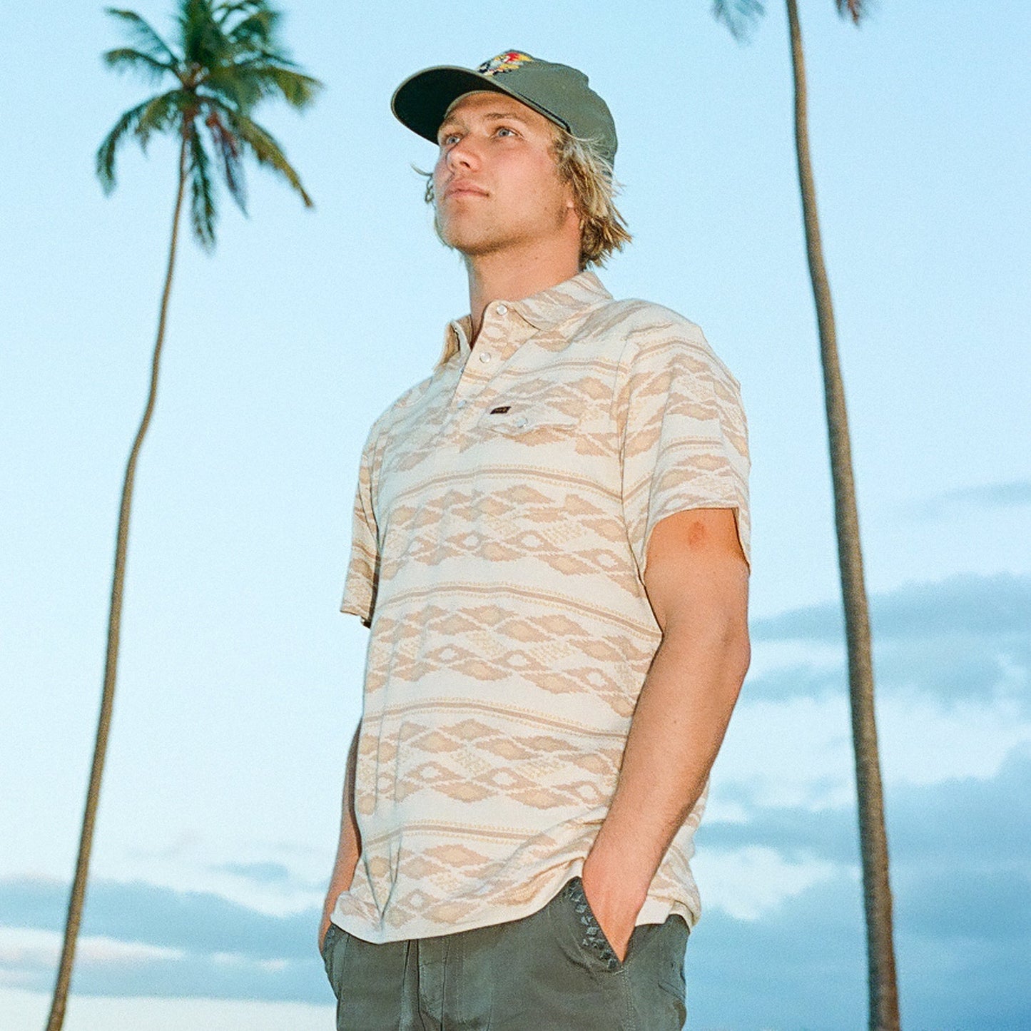 A young man wearing a Ranchero Jacquard Polo stands outdoors with his hands in his pockets. Tall palm trees and a clear sky form the backdrop, highlighting the 100% cotton jacquard pattern of his stylish short-sleeve shirt.