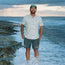 A smiling man stands on a beach with waves around his calves. He wears a lightweight cotton shirt with a single pocket, green cap, and palm tree print shorts. The sky is cloudy and the ocean stretches to the horizon.