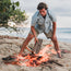 A man in a light blue shirt and cotton twill shorts adds a piece of wood to a campfire on a sandy beach, with the ocean and trees visible in the background.
