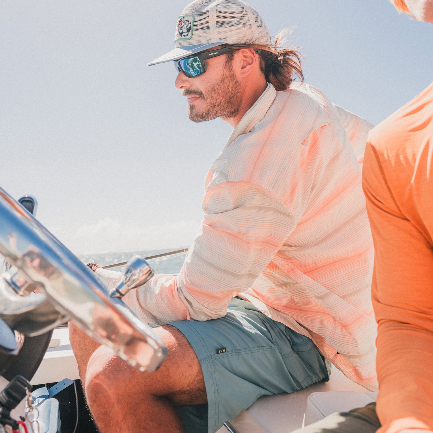 A man wearing sunglasses, a cap, and a light striped shirt steers a boat on a sunny day, sporting versatile Horizon Hybrid Shorts. Another person in an orange shirt is partially visible beside him, capturing the relaxed vibe of the boating trip.