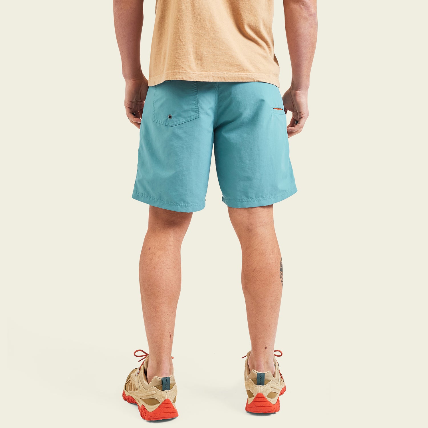 A person stands with their back to the camera, wearing a beige t-shirt, light blue Horizon Hybrid Shorts made from ripstop fabric, and beige athletic shoes with orange soles, set against a plain light background.