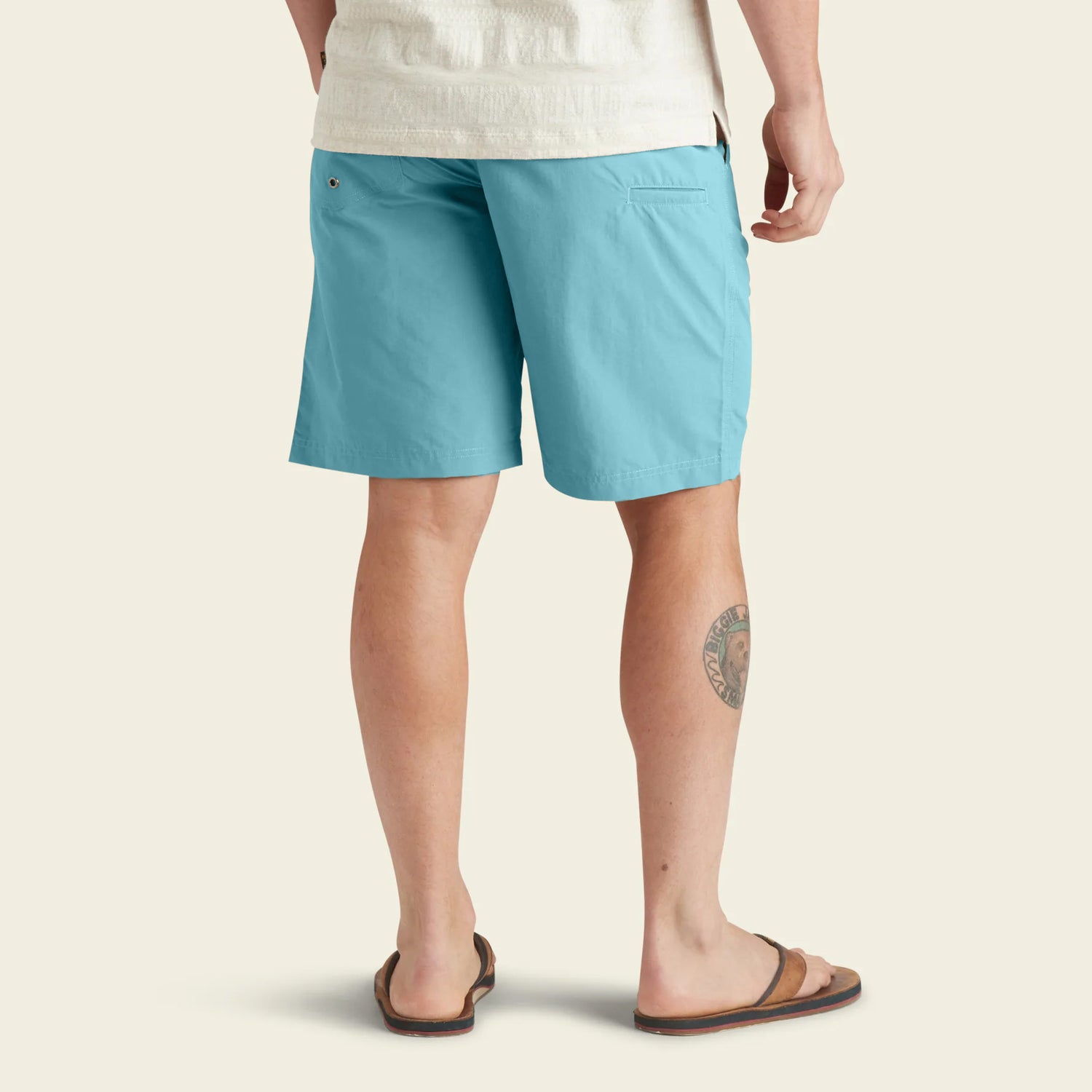 A person wearing light blue Horizon Hybrid Shorts, a light-colored shirt, and brown flip-flops stands facing away, showing a tattoo on their left calf against a plain background.