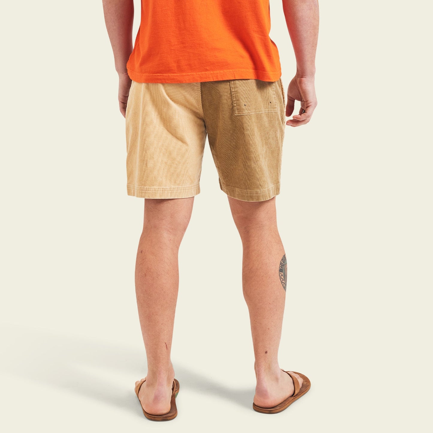 A person wearing an orange T-shirt, Pressure Drop Cord Shorts in color-blocked tan and brown stretch cotton corduroy, and brown flip-flops stands with their back to the camera. A tattoo is visible on their left calf. The background is light beige.