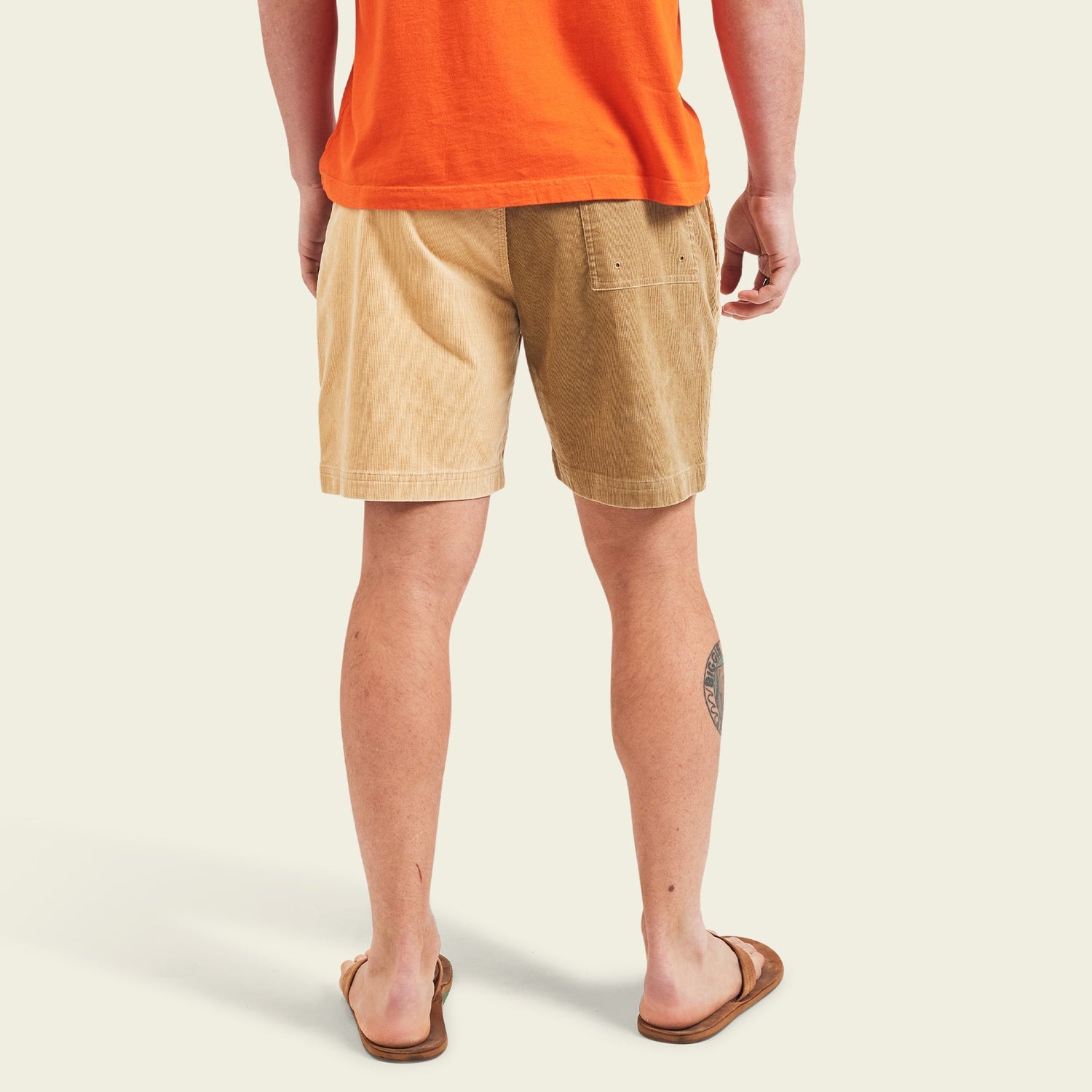 A person wearing an orange T-shirt, Pressure Drop Cord Shorts in color-blocked tan and brown stretch cotton corduroy, and brown flip-flops stands with their back to the camera. A tattoo is visible on their left calf. The background is light beige.