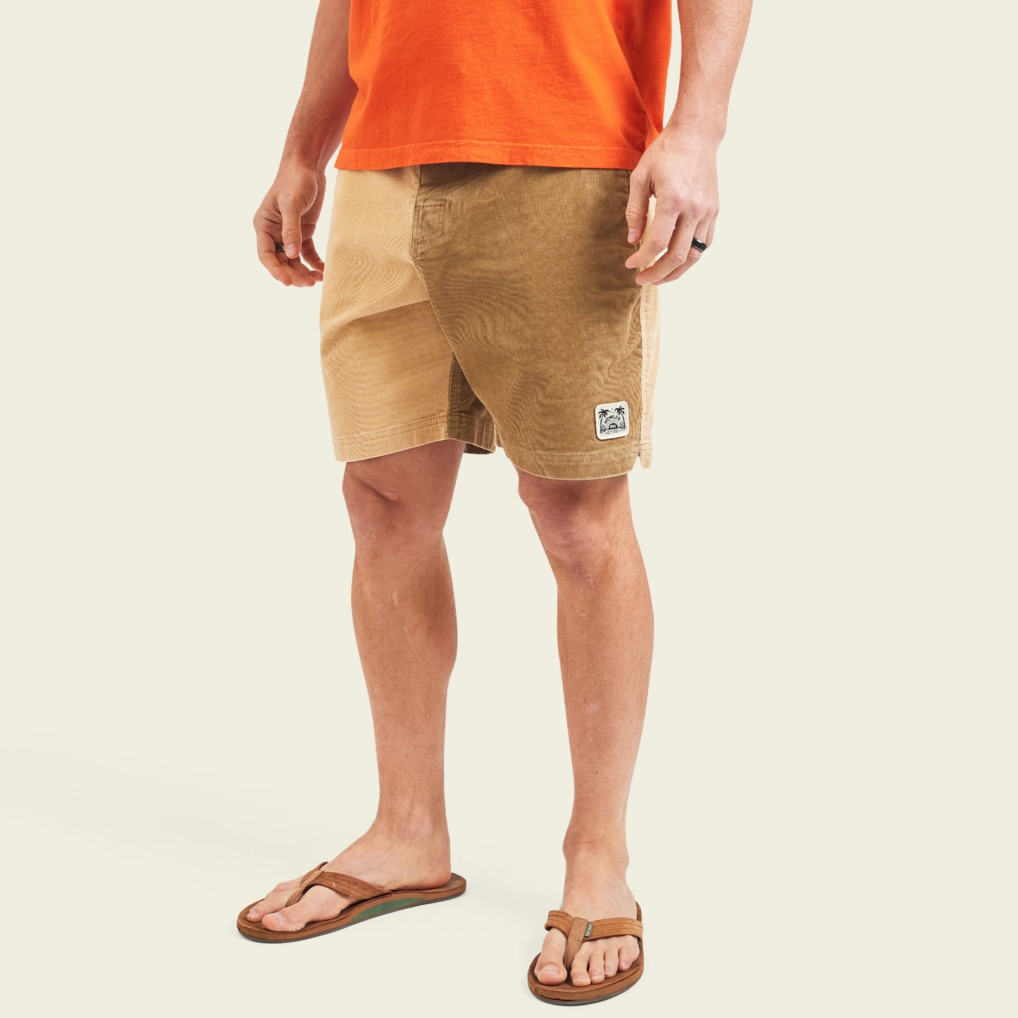 A man wearing an orange T-shirt, Pressure Drop Cord Shorts made from stretch cotton corduroy, brown flip-flops, and a ring on his left hand stands against a plain light background.