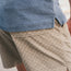 A close-up of a person wearing a blue textured shirt and beige Pressure Drop Cord Shorts crafted from stretch cotton corduroy, with part of their arm visible. The photo highlights the side seam, pocket details, and elastic waistband.