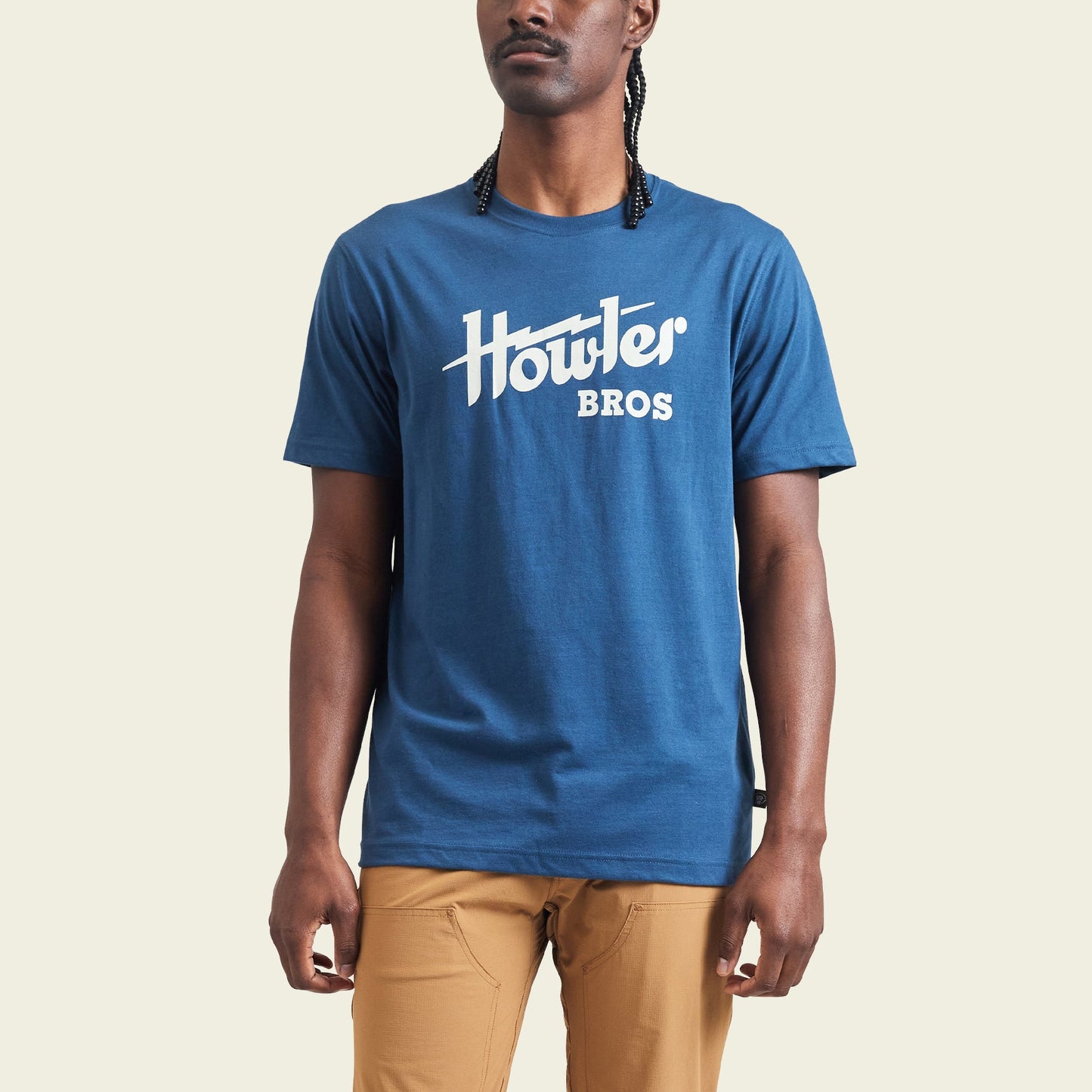 A person wearing a blue cotton-poly blend Howler Bros t-shirt and tan pants stands against a plain, light-colored background. The shirt features a white Howler Bros logo across the chest, showcasing classic Howler designs.