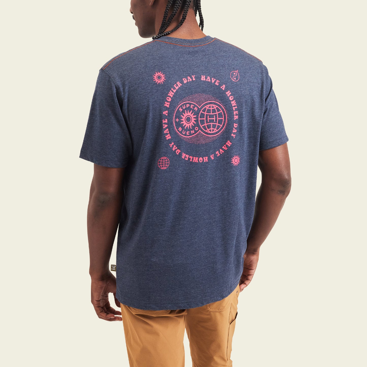 A person wearing a navy blue Howler blended t-shirt with pink graphic designs and text on the back that says Have a dope day in a circle. Made from a soft cotton-poly blend for maximum comfort, paired with khaki pants and facing away to show the design.