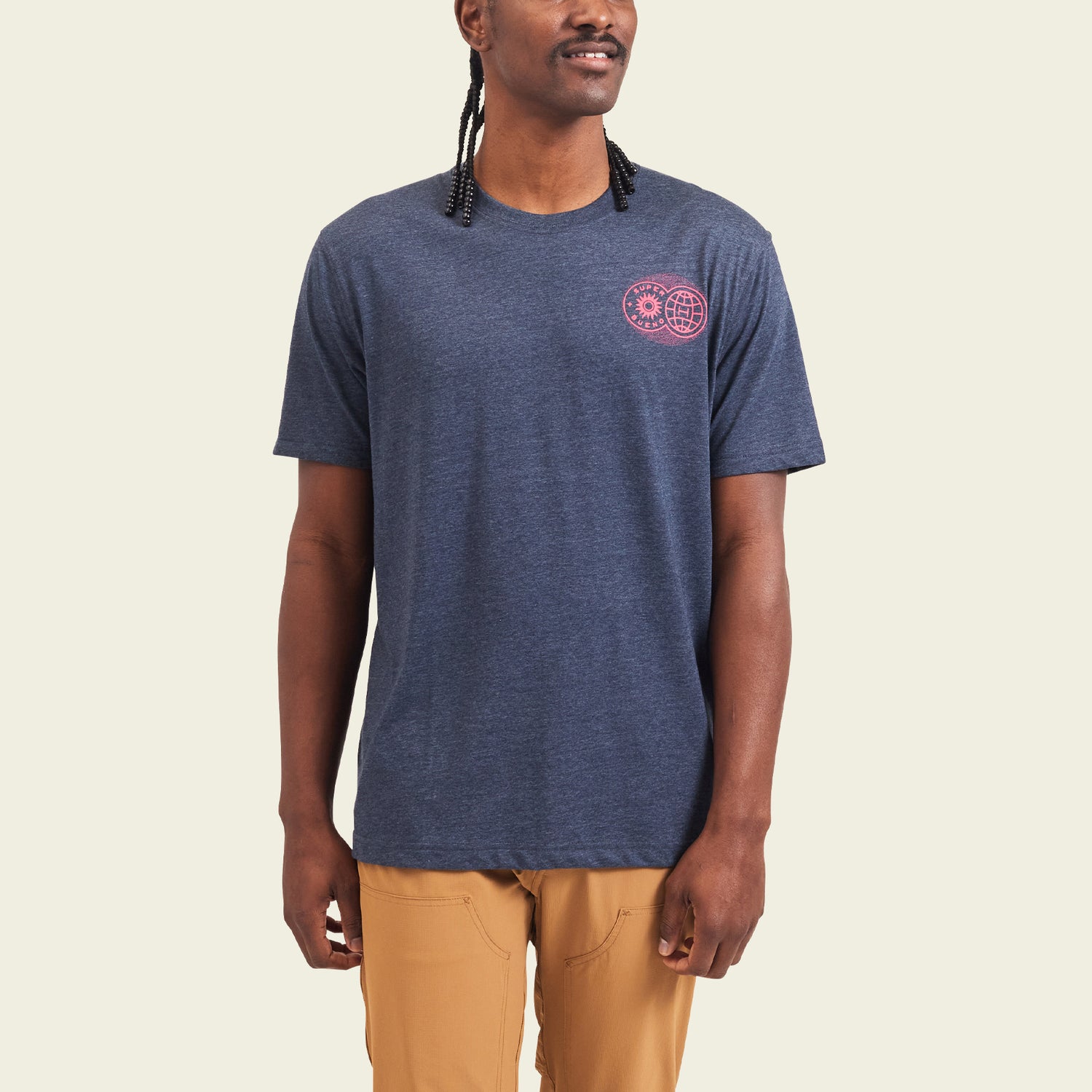 A person wearing a navy blue Howler blended t-shirt with a small pink graphic on the left chest and tan pants stands against a plain light background, enjoying maximum comfort from the soft cotton-poly blend.