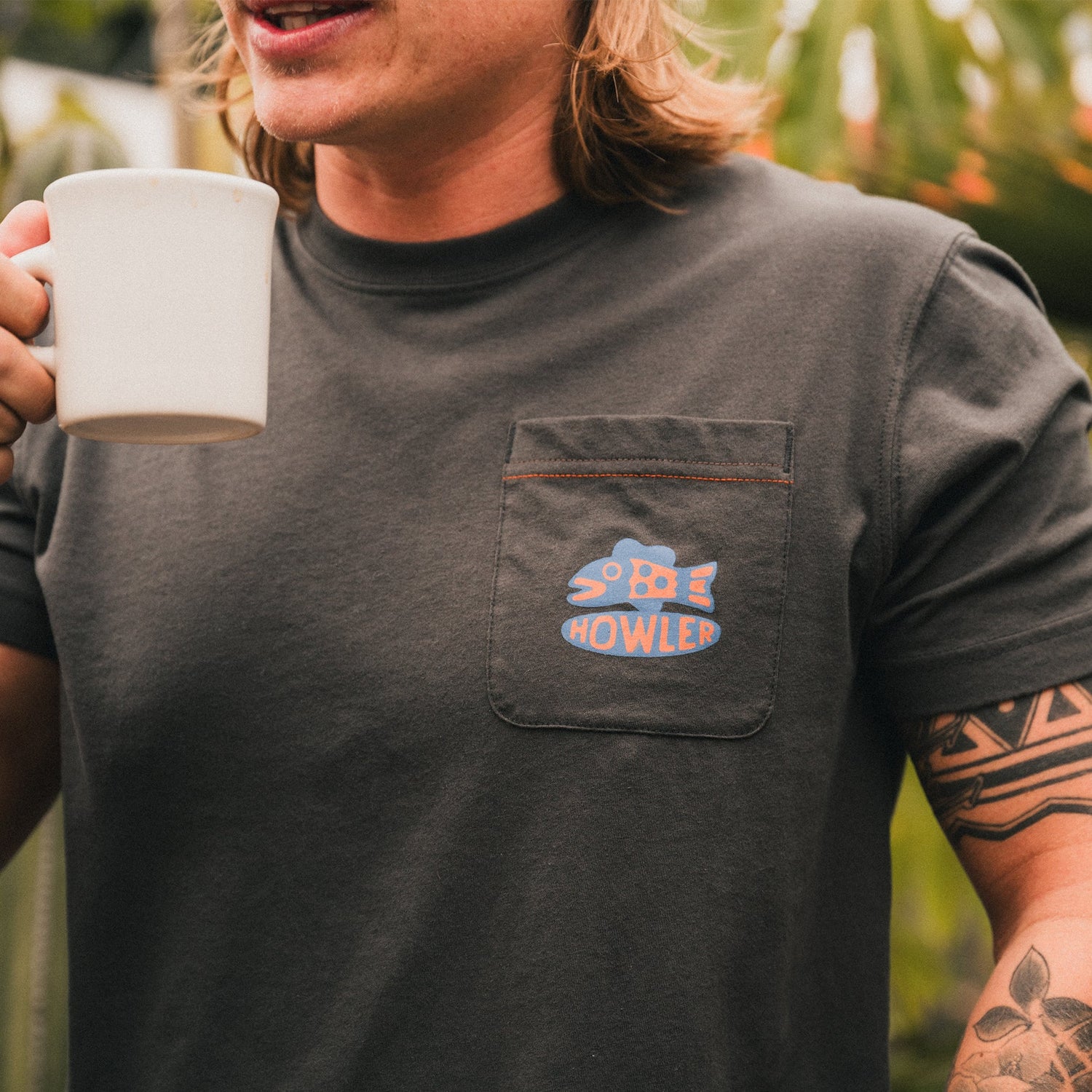 A person with long hair and a partial arm tattoo holds a white mug, wearing a dark Howler blended t-shirt with a pocket featuring the blue and orange Howler logo. Lush greenery is blurred in the background, highlighting maximum comfort.
