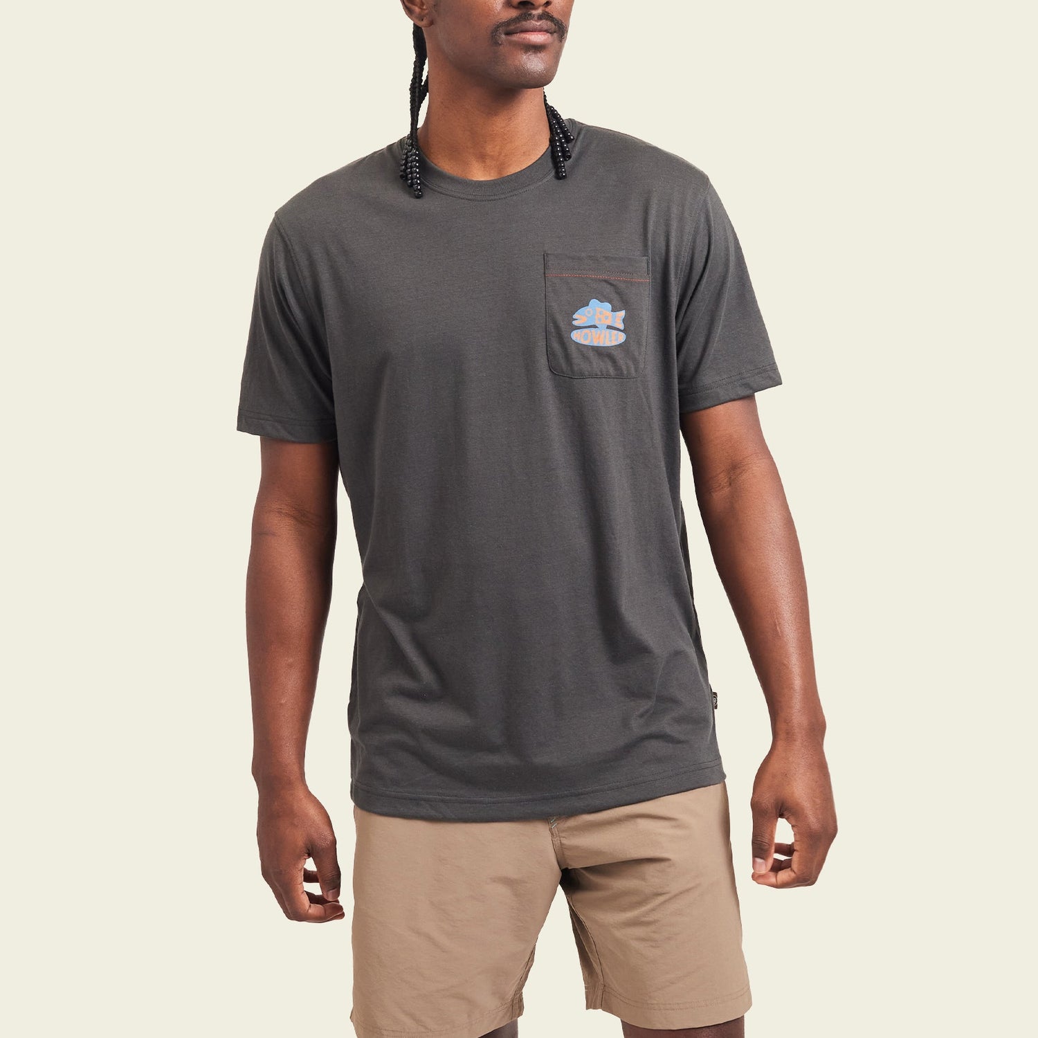 A man wearing a Howler blended t-shirt in charcoal gray, crafted from a cotton-poly blend for maximum comfort, with a small colorful graphic on the chest pocket and paired with light brown shorts, stands against a plain light background.