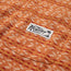 Close-up of an orange patterned cotton jacquard t-shirt with a chest pocket, featuring a white rectangular Howler Brothers label and palm tree graphic. The abstract design in beige and reddish tones evokes a vintage surf shirt vibe.