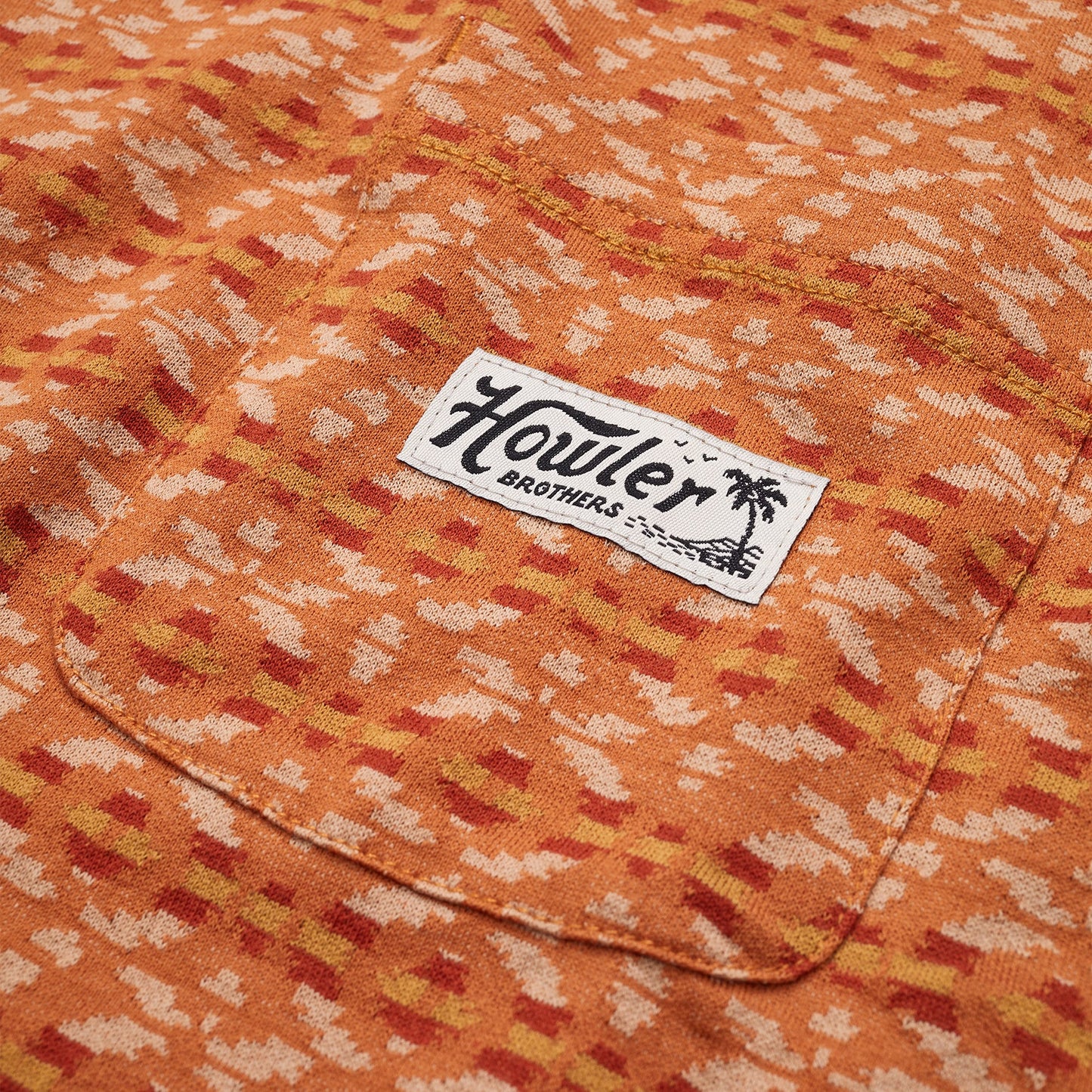 Close-up of an orange patterned cotton jacquard t-shirt with a chest pocket, featuring a white rectangular Howler Brothers label and palm tree graphic. The abstract design in beige and reddish tones evokes a vintage surf shirt vibe.