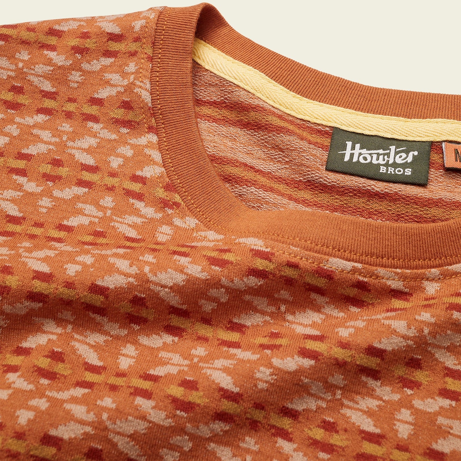 Close-up of an orange and beige patterned Howler Bros sweater, reminiscent of a vintage surf shirt, showing the collar, neck tag, and textured fabric details against a neutral background.