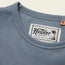 A close-up of a blue heavyweight cotton t-shirt shows the Howler Brothers label with palm tree illustration and the phrase “Heed the Call.” The shirt’s tag indicates a medium size, highlighting its vintage inspired t-shirt charm.