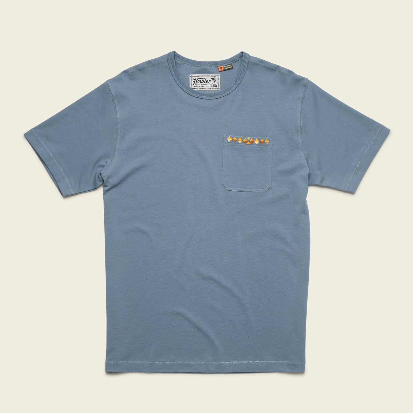 A blue, vintage inspired t-shirt made from heavyweight cotton, featuring a chest pocket adorned with a horizontal row of small, colorful geometric shapes.