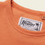 Close-up of an orange heavyweight cotton t-shirt collar with a visible white Howler Brothers label featuring a palm tree illustration and the slogan “Heed the Call.” The shirt’s size “M” tag is partially visible. 80s 90s inspired style.