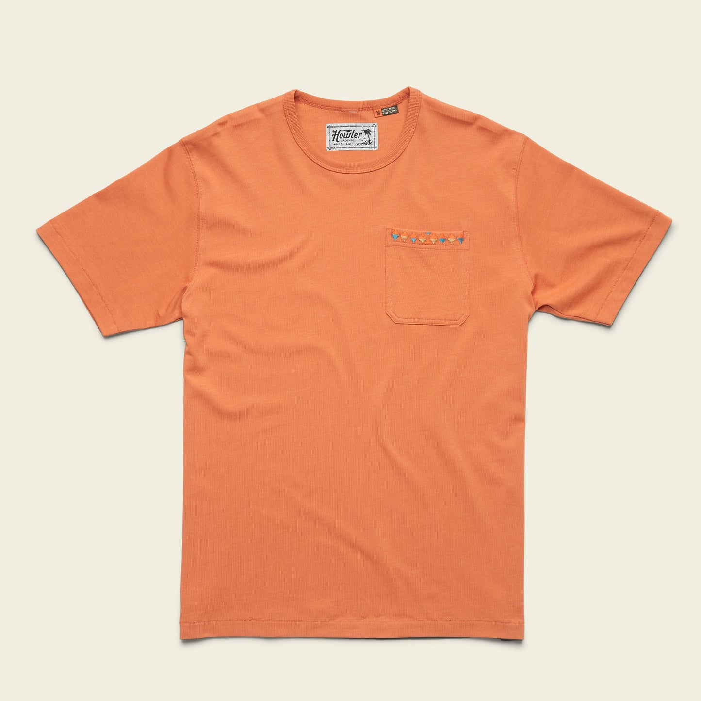An orange heavyweight cotton pocket t-shirt with a chest pocket featuring a small embroidered pattern along the top edge. The shirt is laid flat on a light background, channeling an 80s 90s inspired vibe.