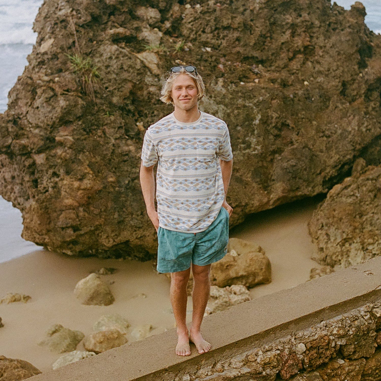 A barefoot person with blond hair, wearing a patterned vintage surfing t-shirt and turquoise shorts, stands on a sandy beach near large rocks, smiling at the camera. Sunglasses rest on their head.