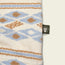 A soft, comfortable terrycloth fabric with a blue, brown, and cream geometric pattern. A small black tag featuring a white monkey face logo is sewn on the right edge of the fabric.
