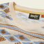 Close-up of a Howler Bros shirt collar, made from comfortable terrycloth with a beige, blue, and brown geometric pattern. A black brand label with white text and a size M orange tag evoke the vibe of vintage surfing t-shirts.