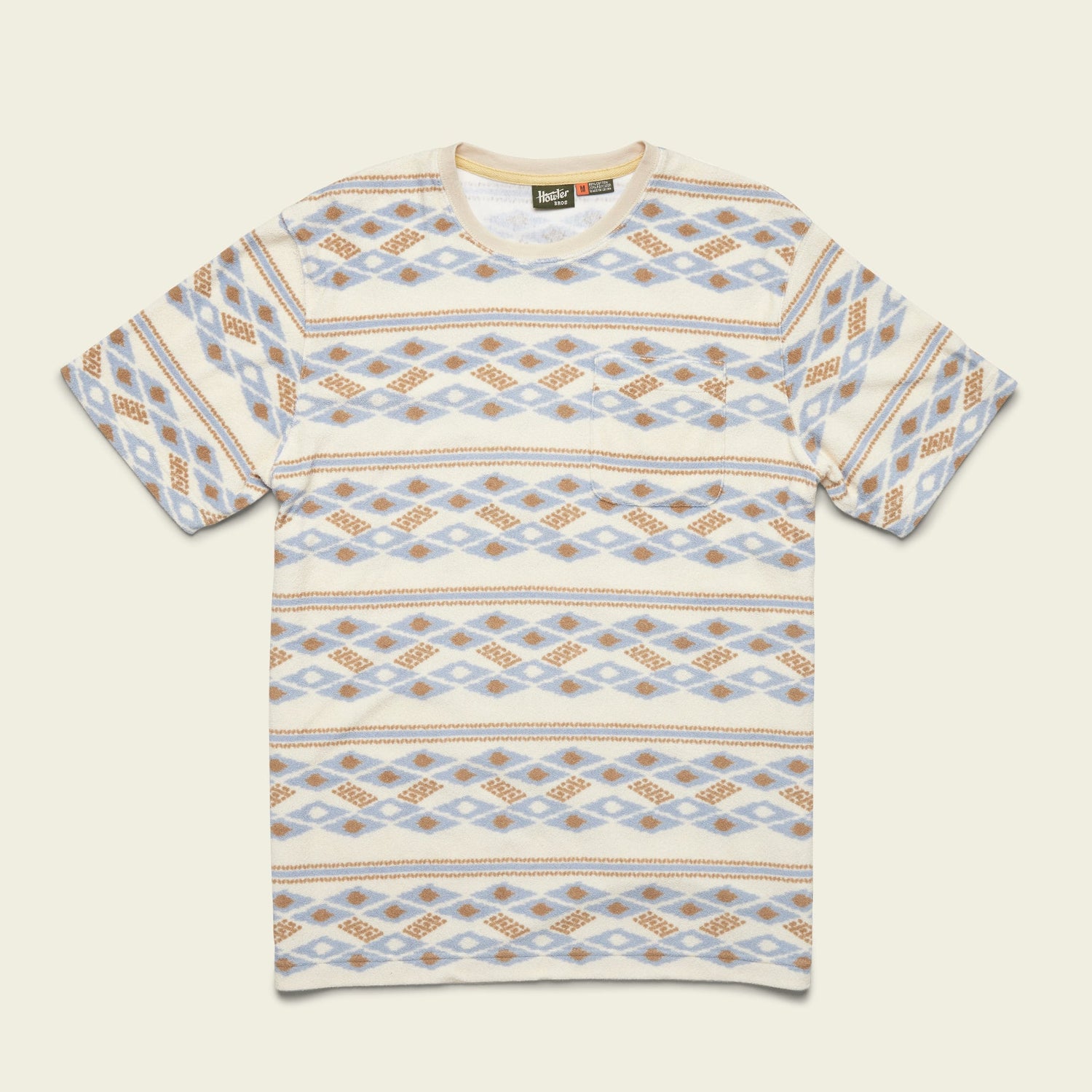 Short-sleeve T-shirt with a crew neck and an all-over geometric pattern in beige, blue, and brown tones arranged in horizontal rows. Inspired by vintage surfing t-shirts and made from comfortable terrycloth, it’s laid flat on a light background.