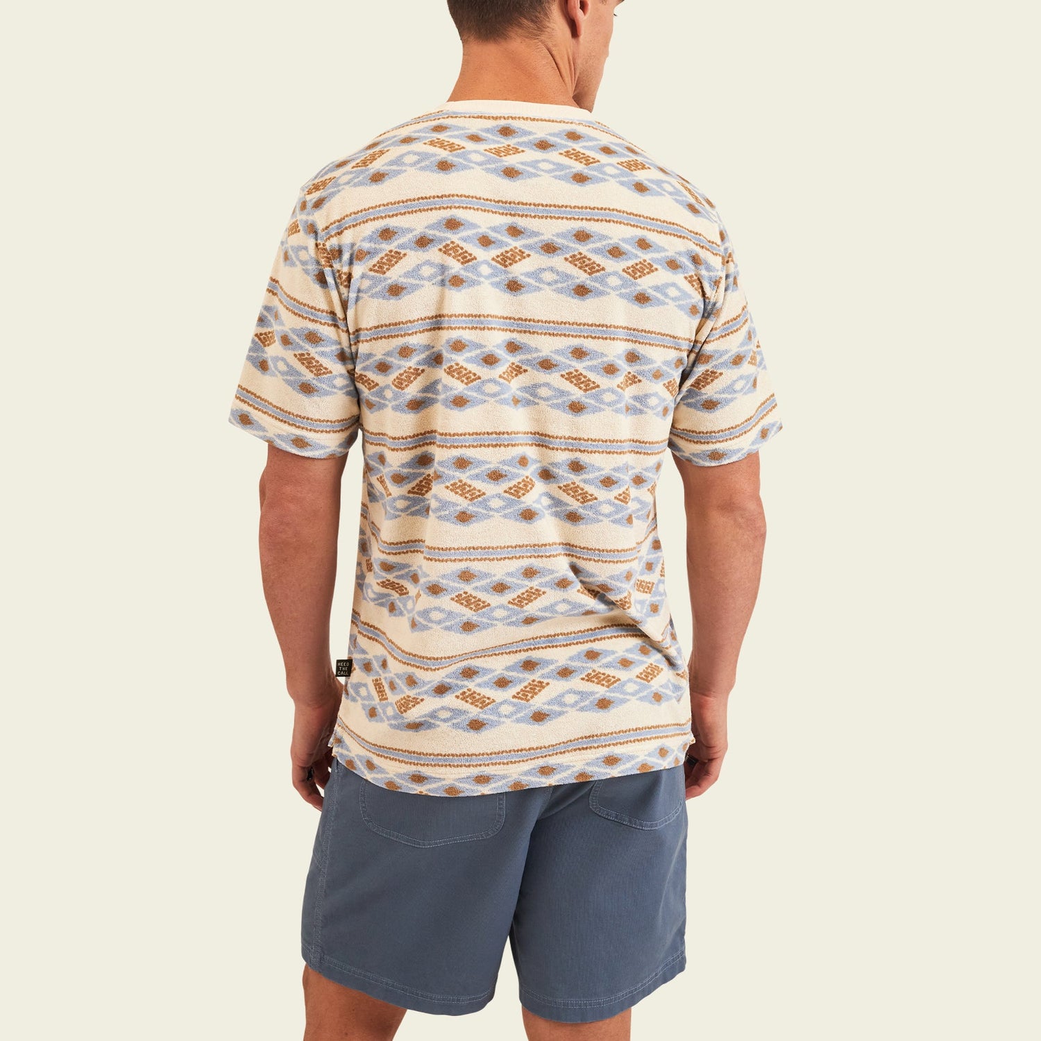 A man stands facing away, wearing a comfortable terrycloth short-sleeve shirt with blue and brown diamond designs and light blue shorts against a plain light background.