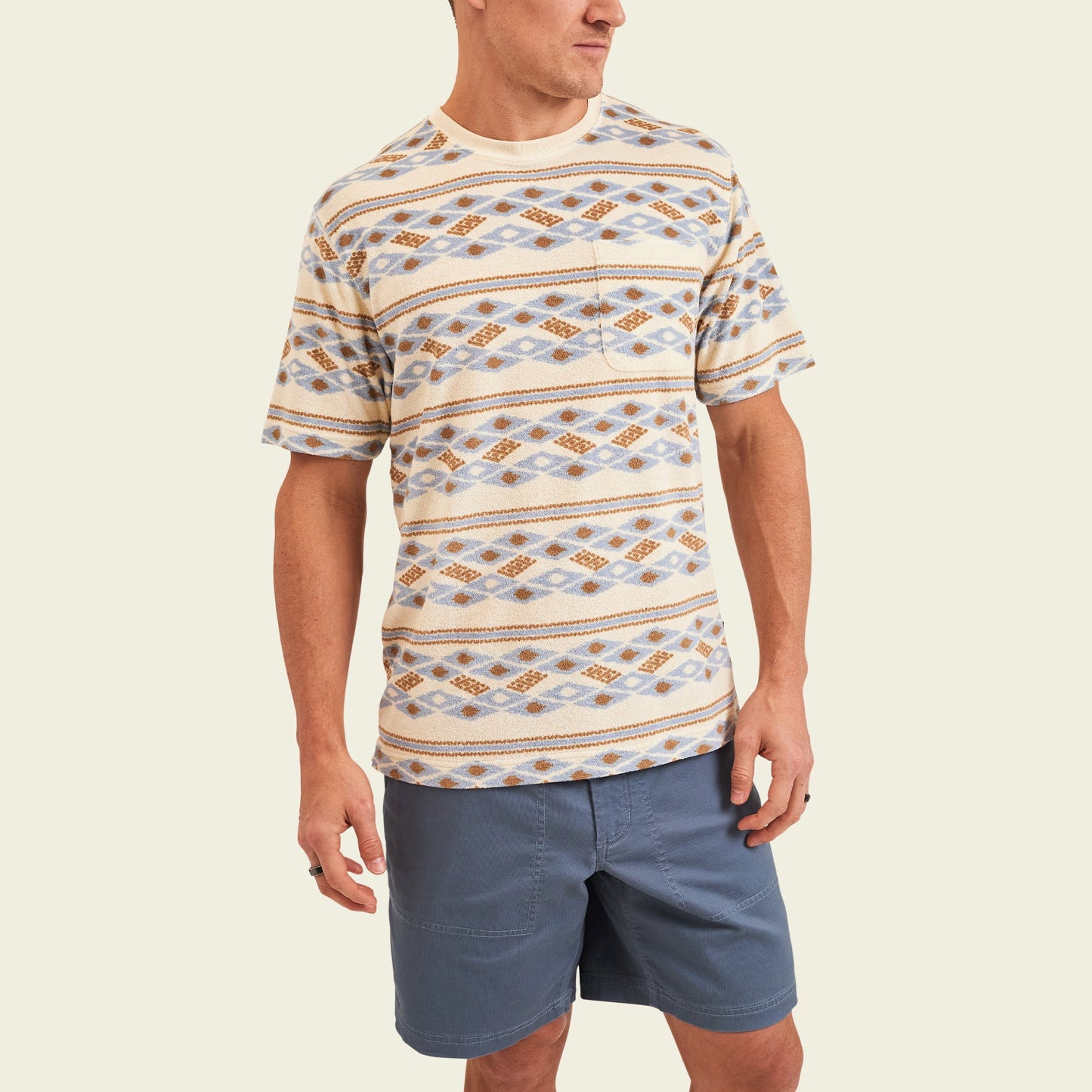 A man wearing a patterned short-sleeve beige and blue diamond print shirt, inspired by vintage surfing t-shirts, and blue shorts stands against an off-white background, looking slightly to his left.
