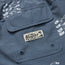 Close-up of a blue fabric pocket with a Howler Brothers label sewn on, seen on their boardshorts. The fabric features a white bird pattern and two metal eyelets at the bottom of the pocket.
